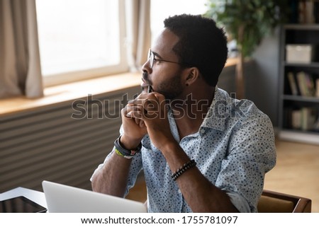 African guy sitting at table near laptop pondering over problem search answer looking at distance out the window. Student or office worker having doubts, feel uncertain thinking about solution concept Royalty-Free Stock Photo #1755781097