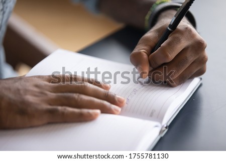 African man sit at desk hold pen keeps startup business ideas, plans, creative thoughts to notebook close up image. Makes appointment notes time, writes important things and to-do list not to forget Royalty-Free Stock Photo #1755781130