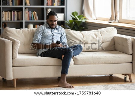 Lazy african man in casual clothes barefoot spend time in modern warm living room holds smart phone chatting with friend, read media news, play on-line game, gambling, gadget overuse addiction concept