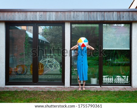 A pretty girl in a summer dress poses against the background of the pool window in the backyard with an inflatable ball.