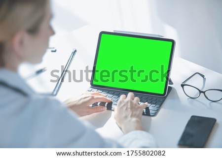 Medical worker a woman in a white coat uses a tablet in the office at the Desk, chromakey on the tablet screen, a view over her shoulder