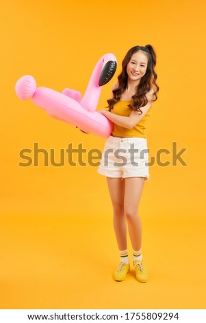 Happy young Asian woman dress casual clothes posing with inflatable flamingo over orange background.