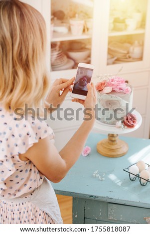 Housewife texing, taking picture of sweet cake she baked. In her kitchen at home. She's wearing apron over polka dot dress. From behind.