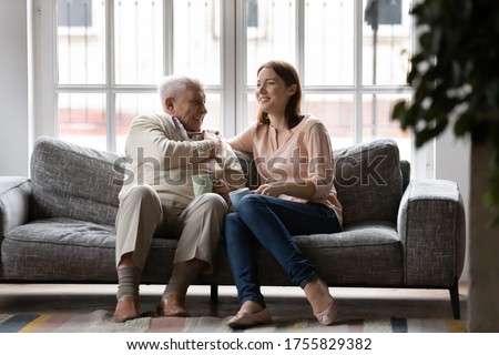 Happy older man and adult daughter drinking tea or coffee together, sitting on cozy couch, smiling young woman and mature father having fun, enjoying leisure time, talking, sharing news Royalty-Free Stock Photo #1755829382