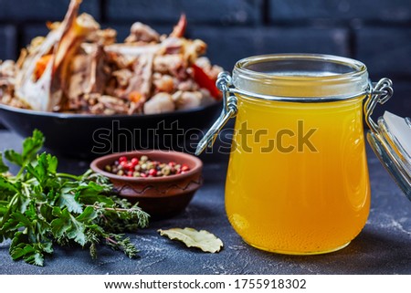 cooked chicken stock in a glass jar on a kitchen table with bouquet garni and chicken bones in a bowl on a background of a brick wall, horizontal view from above, close-up