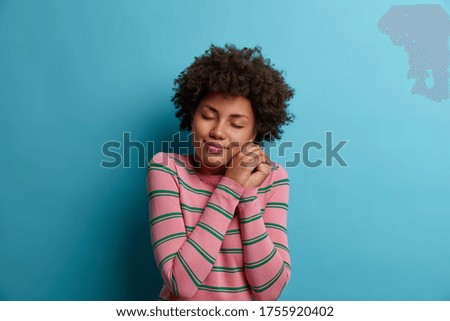 Beautiful dark haired woman poses with closed eyes, keeps hands near face, imagines something pleasant, enjoys peaceful domestic atmosphere, wears pink striped jumper, models over blue background