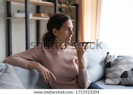 Smiling young woman recording voice message, holding phone near face, sitting on cozy couch at home, chatting online, making call, talking by speakerphone, activating assistant on smartphone