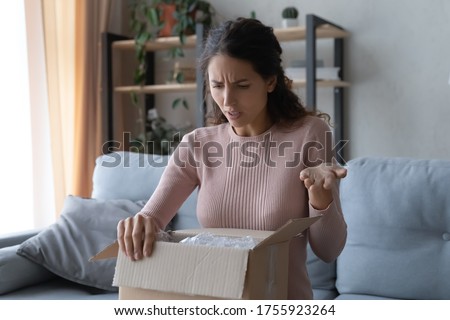 Angry confused woman unpacking parcel, wrong or broken online store order, sitting on couch at home, dissatisfied female looking in cardboard box, bad delivery service, displeased by post shipping Royalty-Free Stock Photo #1755923264