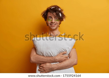 Positive Caucasian adult man awakes early in morning, has good mood, wears under eye patches for reducing puffiness, embraces soft pillow, isolated on yellow background. Morning time concept