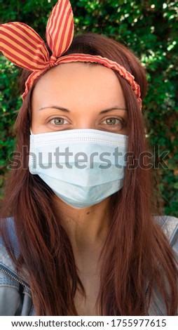 Close up portrait of a Caucasian woman with a medical mask and a striped headband outside in nature looking at the camera.