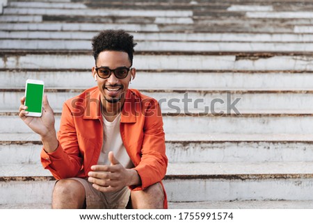 Young happy curly brunet man in orange jacket, beige shorts and sunglasses sits on stairs outside and demonstrates phone screen.