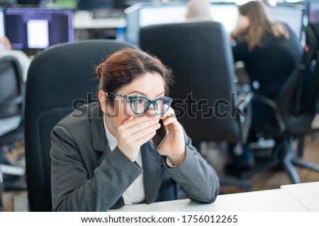 A woman sits in an office at her desk and gossips on the phone. Corporate ethics. Female employee in a suit tells secrets on a smartphone at work. Royalty-Free Stock Photo #1756012265
