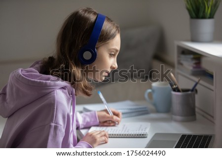 Concentrated teenage girl in wireless headset sit at desk at home studying on laptop, focused teenager do prepare homework handwrite in notebook, using modern computer, distant education concept