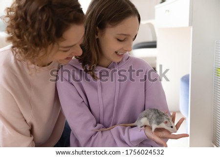 Happy mother and teenage daughter enjoy playing with cute domesticated mouse at home together, loving smiling mom and teen girl child have fun take care of small domestic rat animal or pet
