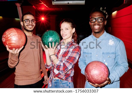 Pretty young woman and two intercultural guys with toothy smiles holding bowling balls while playing together in entertainment club