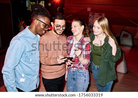 Young smiling man with smartphone showing friends photo of his girlfriend taken by him during game of bowling in leisure center