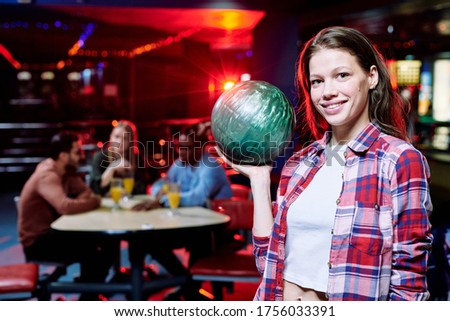 Pretty smiling girl in casualwear holding bowling ball while standing in front of camera on background of group of intercultural friends