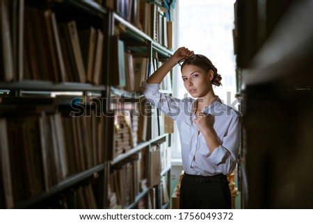 Beautiful female student in a white shirt stands between the rows in the library, bookshelves worth of books. Dark photo