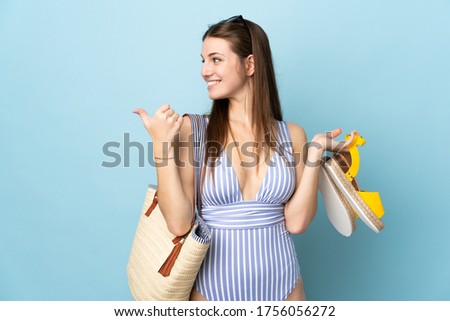 Young caucasian woman holding a beach bag an summer sandals isolated on blue background pointing to the side to present a product