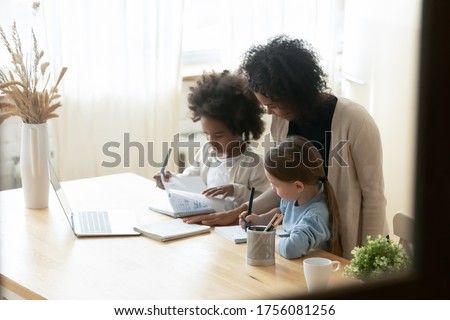 African mom of two multiethnic school age daughters do lesson homework helps them. American and Caucasian girls siblings writing on workbooks study with teacher at home. Homeschooling tutoring concept Royalty-Free Stock Photo #1756081256