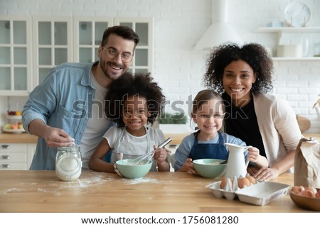 Multiethnic family with kids engaged in holiday cake preparation using fresh products mixing eggs milk flour ingredient looking at camera. Teach children, happy parenthood, common hobby, unity concept
