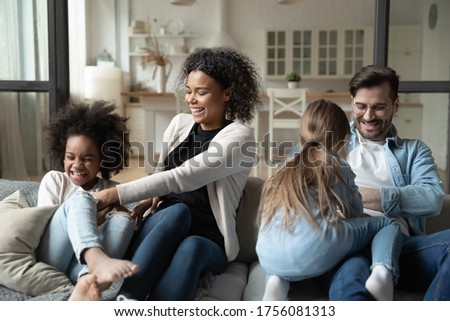 African mom Caucasian dad play with multi-racial daughters tickling girls laughing feels good, family spend funny time seated on couch in living room enjoy weekend. Happy homeowners having fun concept Royalty-Free Stock Photo #1756081313