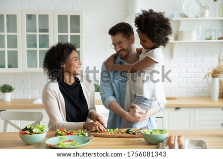 Caucasian dad holds on hands little mixed-race daughter while African wife preparing healthy vegetable salad family enjoy conversation in kitchen. Healthy home food, communication and cookery concept Royalty-Free Stock Photo #1756081343