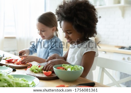 African and Caucasian little girls best friends cooking together in modern kitchen. Multiracial cousins hold knives cutting vegetables on wooden board prepare healthy salad making surprise for parents Royalty-Free Stock Photo #1756081361