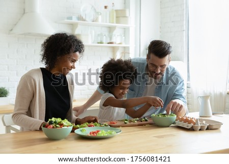 Caring multiethnic couple teach small daughter cook vegetable salad in modern domestic kitchen. Family enjoy cooking and communication together, healthy organic food preparation, love and bond concept