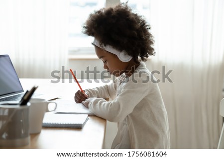 Side view small African girl wear wireless headphones doing homework seated at desk in her room. Focused schoolgirl listen audio lesson use laptop writes on workbook. E-learning, homeschooling concept Royalty-Free Stock Photo #1756081604