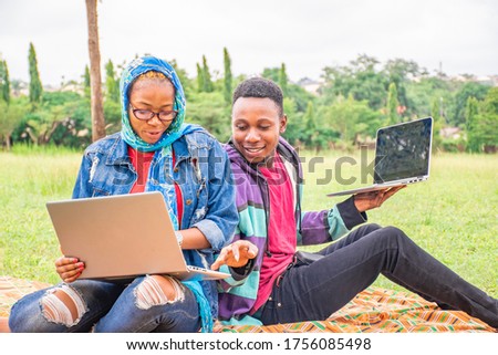 two young black people, sitting outdoors in a park, working with laptop computers together on a project