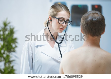 A friendly young doctor in a white coat examines a male patient using a stethoscope in a bright office. Medical consulting service. Concept of medicine and healthcare.