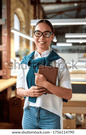 Portrait of attractive ambitious asian businesswoman in casual wear and glasses holding notebook, while smiling at camera. Modern working space in the background. Vertical shot. Front view