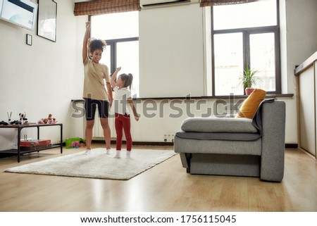 Full length shot of african american woman baby sitter and caucasian cute little girl having workout at home. Children education, leisure activities, babysitting concept. Horizontal shot