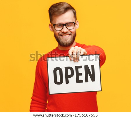Happy bearded man smiling for camera and showing   signboard with Open inscription against yellow background
