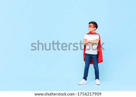 Superhero boy in red mask and cape with arms crossed thinking and looking at copy space aside isolated light blue background