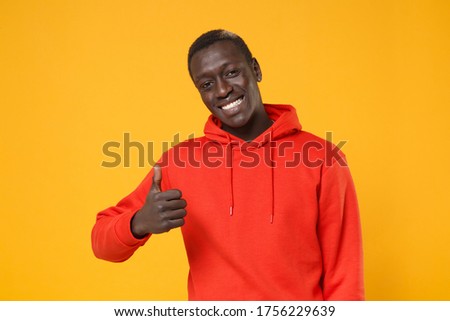 Cheerful young african american man guy in red streetwear hoodie posing isolated on yellow background studio portrait. People sincere emotions, lifestyle concept. Mock up copy space. Showing thumb up