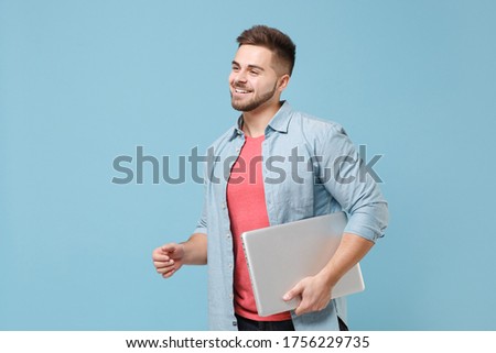 Smiling young bearded guy 20s in casual shirt posing isolated on pastel blue background studio portrait. People emotions lifestyle concept. Mock up copy space. Hold laptop pc computer, looking aside