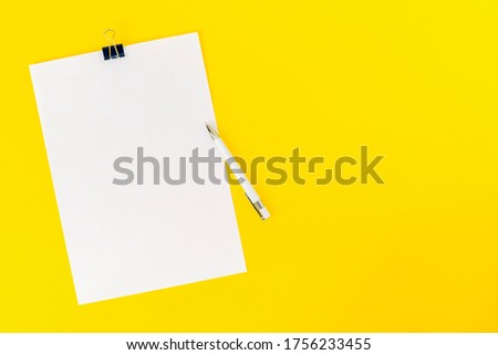 An empty sheet of white office paper is fastened with a stationery clip and a pen on a yellow background. Mockup, blank for board announcement, information, statement, school. Top view, copy space.