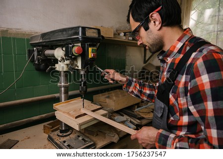 Working process in the carpentry workshop.A man in overalls works in a carpentry workshop.A man drills wooden board with a drilling machine.Profession, carpentry, woodwork and people concept Royalty-Free Stock Photo #1756237547