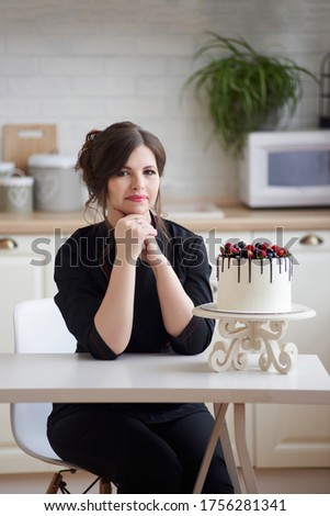 Confectioner young woman happy with her work sitting at table clasped hands looking at camera with beautiful juicy berry cake. Employment, freelancer at home concept, copy space.