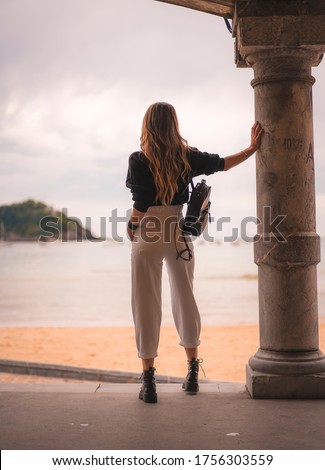 Lifestyle in the city with a blonde girl in white pants and a leather jacket near the beach. Photos next to a column looking at the sea from behind on a sunset