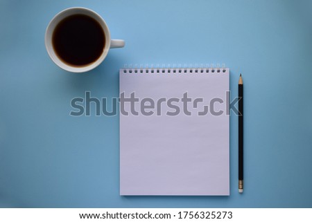 A white Notepad with a black pencil and a white coffee Cup on a blue background. Desktop, workspace. The view from the top.