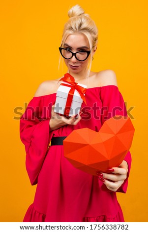 European girl in a red dress holds a gift and a red heart on an orange studio background