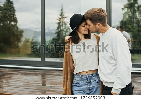 Stylish hipster couple hugging on wooden porch, relaxing in modern cabin in mountains. Happy young family in modern outfits embracing on background of terrace and big window