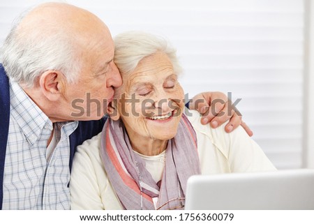 Happy senior kisses his smiling wife on the cheek at the computer