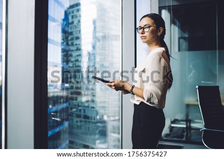 Pensive businesswoman dressed in formal apparel standing near panoramic window in enterprise company and thinking about trade investment,thoughtful female entrepreneur in eyewear for vision protection Royalty-Free Stock Photo #1756375427