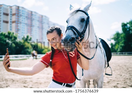 Caucasian female jockey enjoying free time for horseback in paddock country club and clicking selfie pictures for sharing content to social networks, millennial making image with friendly stallion