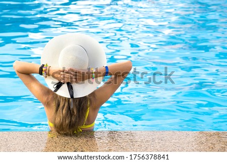 Back view of young woman with long hair wearing yellow straw hat relaxing in warm summer swimming pool with blue water on a sunny day.