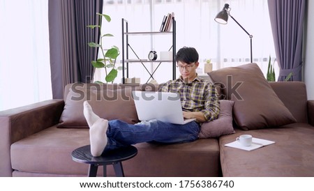 handsome asian freelancer working on his laptop computer remotely from home, sitting on the cozy beige sofa at home, wearing casual outfit